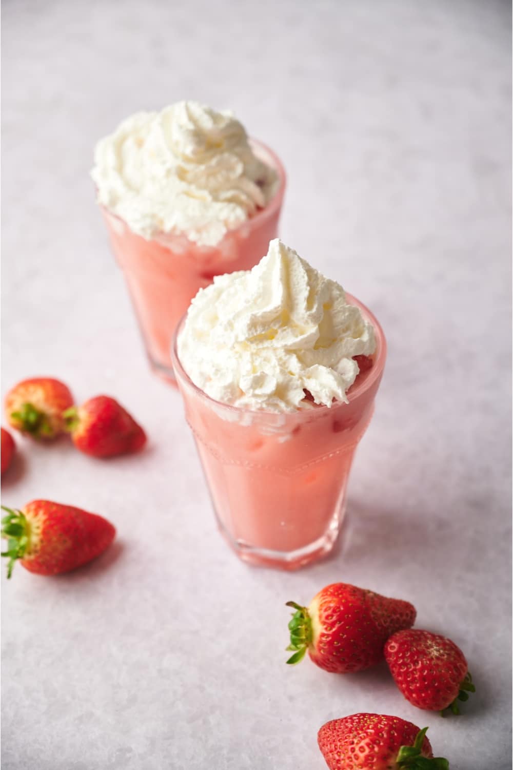 Two glasses that are filled with a pink drink with whipped cream on top. They're on a grey counter with strawberries on it.