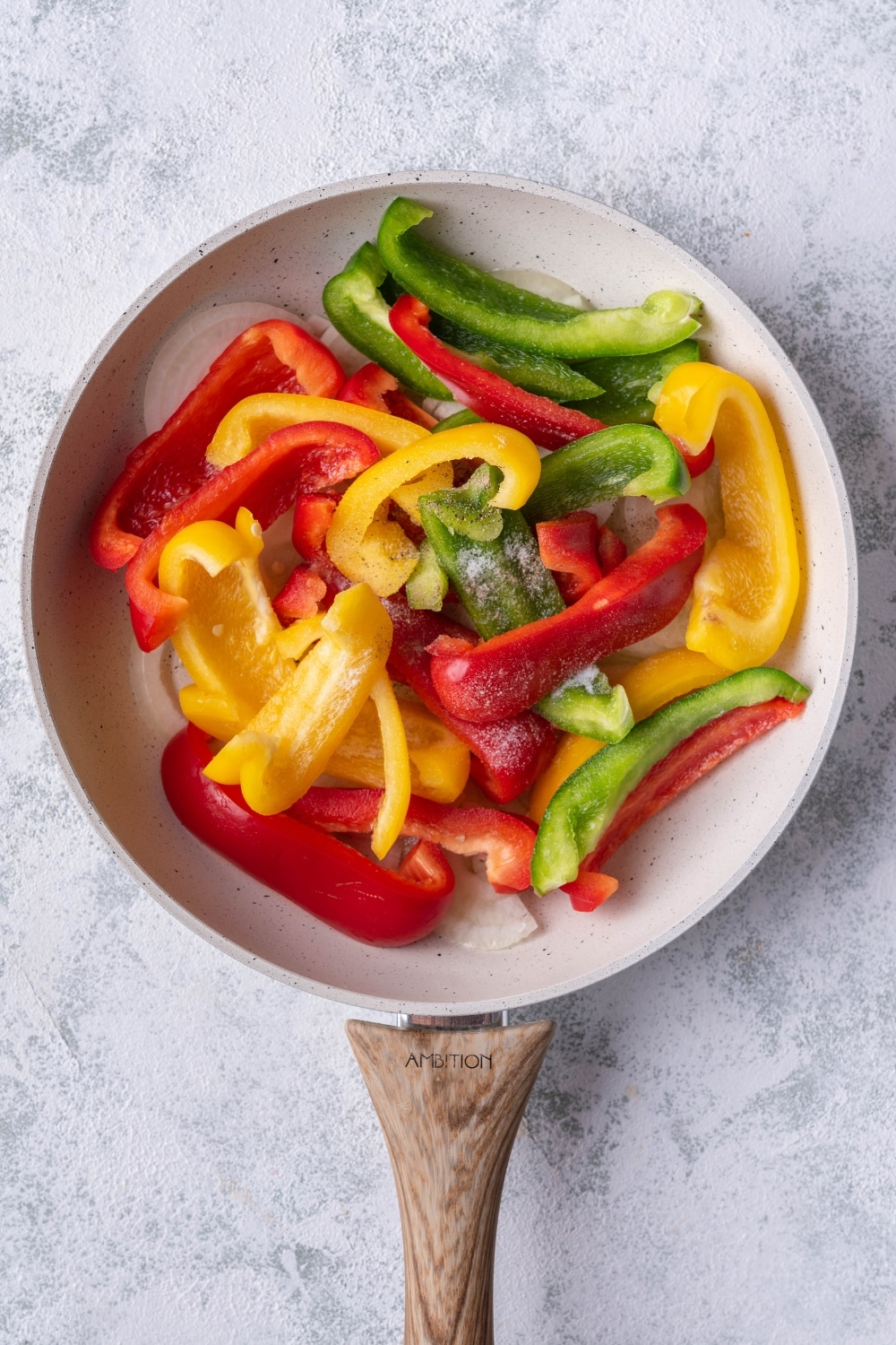 Uncooked sliced bell peppers and onions in a white speckled skillet with a wooden handle. The skillet is resting on a grey countertop.