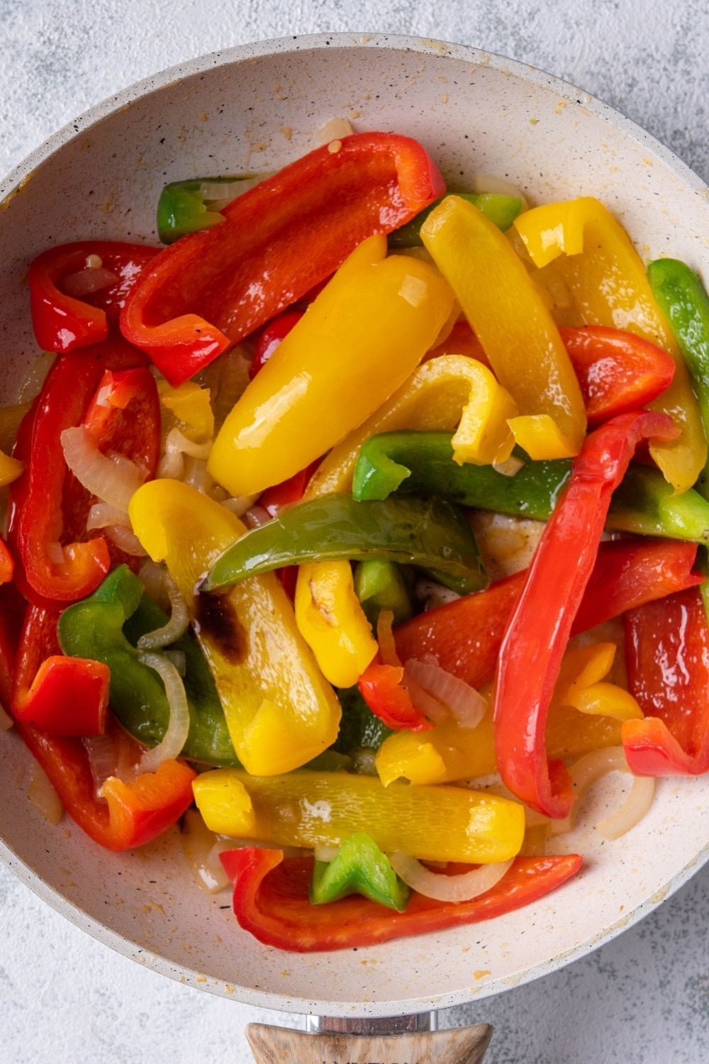 Close up of sauteed bell peppers and onions in a white speckled skillet with a wooden handle. The skillet is resting on a grey countertop.