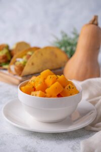 Sauteed butternut squash cubes garnished with herbs in a white bowl set on top of a white plate. Behind is a plate of hard shell tacos topped with butternut squash cubes next to a whole butternut squash.