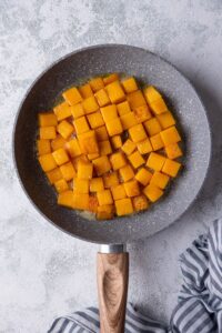 Sauteed butternut squash cubes in a grey speckled pan with a wooden handle. The pan is resting on a grey countertop.