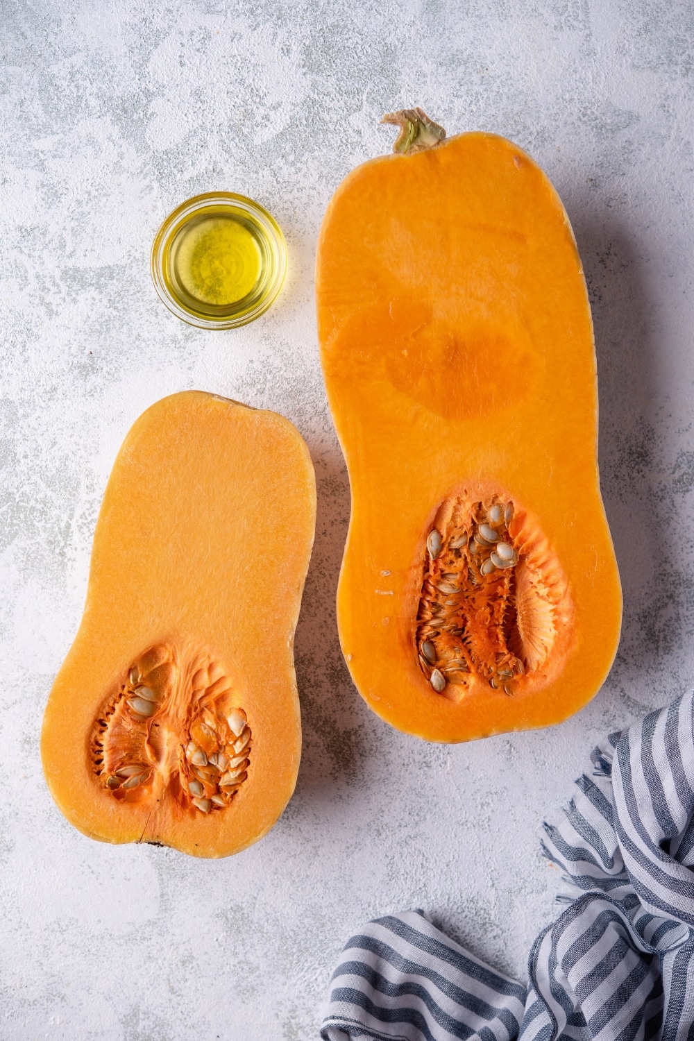 Two cut halves of a butternut squash, the one on the right is slightly bigger and with a deeper orange hue than the one on the left, and a small glass bowl of olive oil.