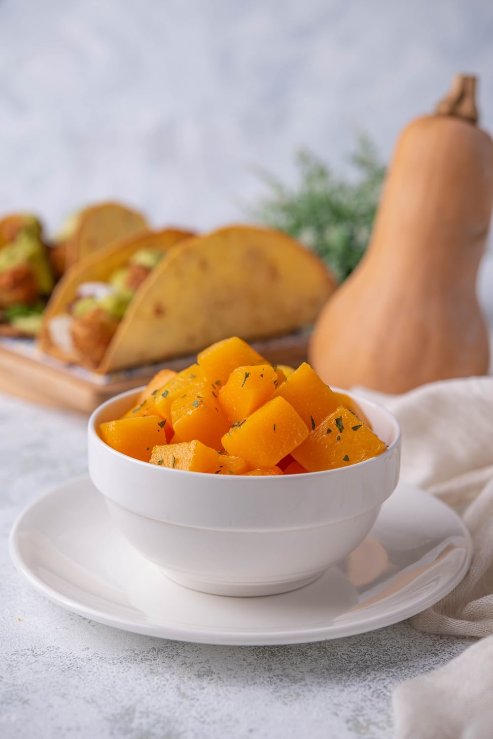 Sauteed butternut squash cubes garnished with herbs in a white bowl set on top of a white plate. Behind is a plate of hard shell tacos topped with butternut squash cubes next to a whole butternut squash.