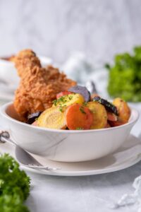 Sauteed carrots and breaded fried chicken drumsticks in a white bowl. The bowl is on a white plate with a fork. Bunches of fresh parsley are laid out on the table.
