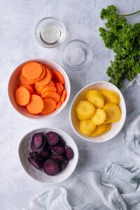 White bowls filled with slices of violet carrots, orange carrots, and yellow carrots, small glass bowls of sea salt and olive oil, a bunch of fresh parsley, and a tea towel.