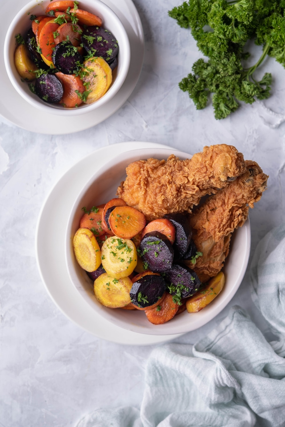 Top view of sauteed carrots and breaded fried chicken drumsticks in a white bowl over a white plate. A second bowl of sauteed carrots garnished with parsley is on the side.