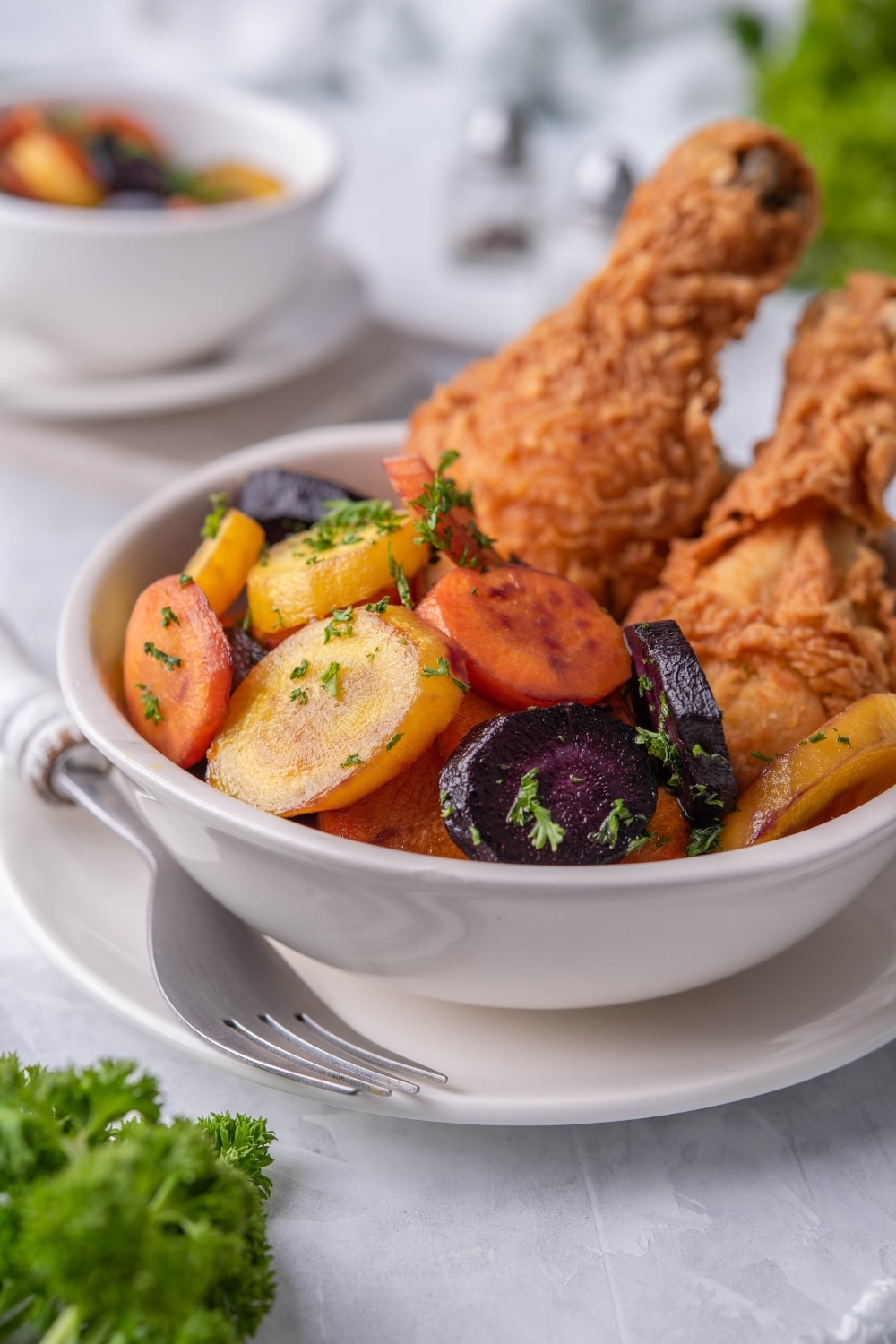 Close up of sauteed carrots in a white bowl with a pair of breaded chicken drumsticks. A second bowl of sauteed carrots can be seen in the back.