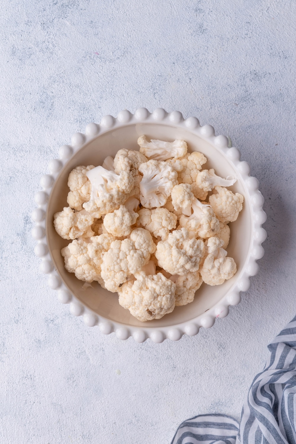 Uncooked cauliflower florets in a white bowl. A striped tea towel is on the side.