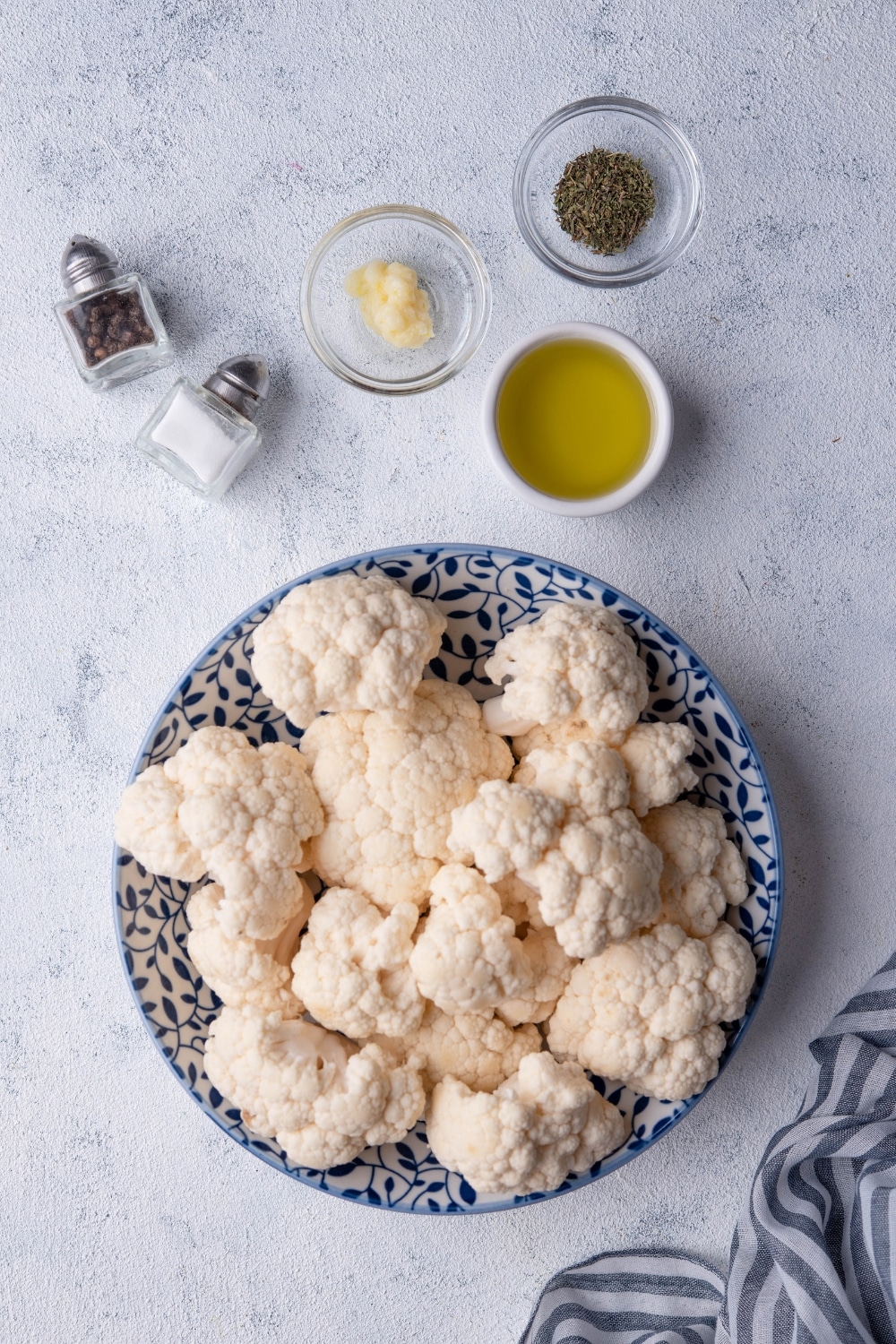 Raw cauliflower in a white bowl decorated with blue leaves, a small white bowl of olive oil, small glass bowls of garlic paste and dried thyme, and salt and pepper shakers.