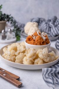 Sauteed cauliflower on a white plate. On the same plate is a small bowl of fried chicken drizzled with white sauce. In the back is a plate of raw cauliflower with a tea towel draped to the side and a pair of salt and pepper shakers.