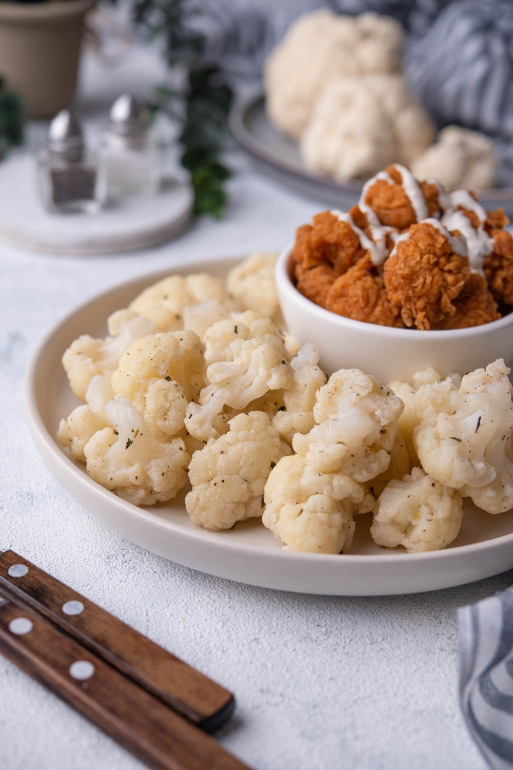 Close up of sauteed cauliflower on a white plate with a small bowl of fried chicken drizzled with white sauce. Part of a pair of utensils with wooden handles is in front of the plate and In the back is a plate of raw cauliflower and a pair of salt and pepper shakers.