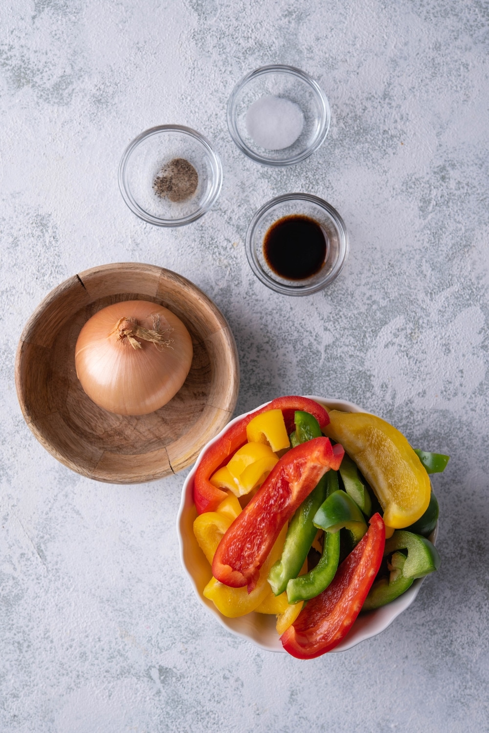 A white bowl of sliced red, green, and yellow bell peppers, a whole onion in a medium wooden bowl, and small glass bowls of salt, pepper, and balsamic vinegar.