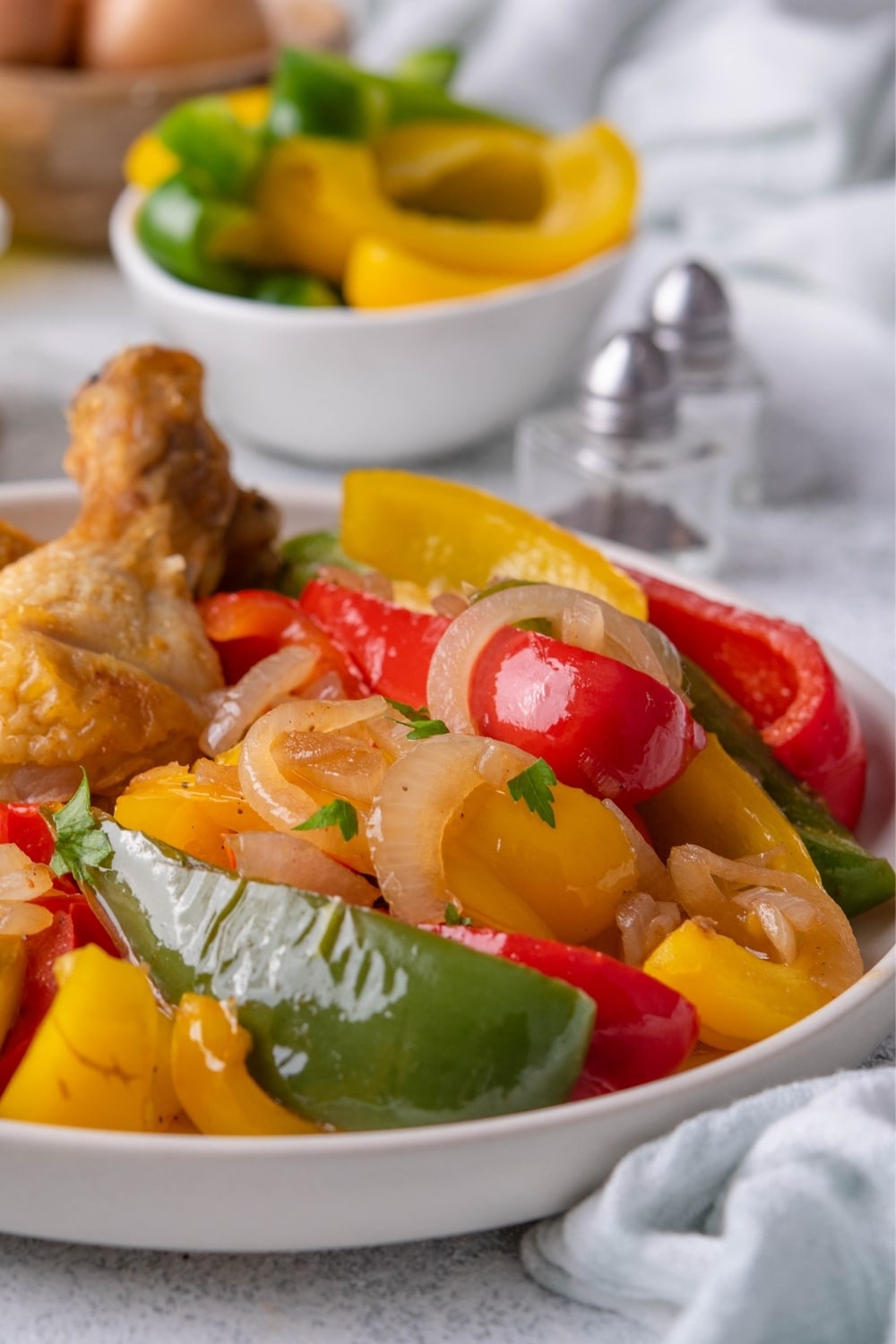 A closer look at sauteed bell peppers and onions garnished with fresh parsley on a white plate. Part of a piece of fried chicken can be seen on the side on the same plate. Behind are salt and pepper shakers and a bowl of raw sliced bell peppers.