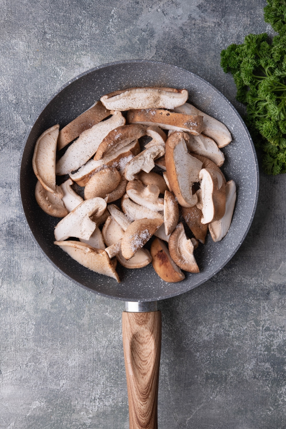 Raw sliced shiitake mushrooms in a grey speckled pan with a wooden handle. The pan is on a grey countertop next to a bunch of fresh parsley.