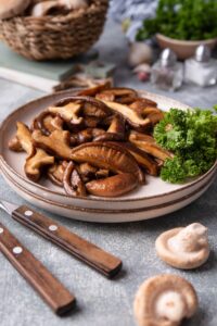 A plate of sauteed shiitake mushrooms with curly parsley on the side. Part of a fork and knife set are beside it and behind is a basket of fresh mushrooms and a small bowl of parsley.