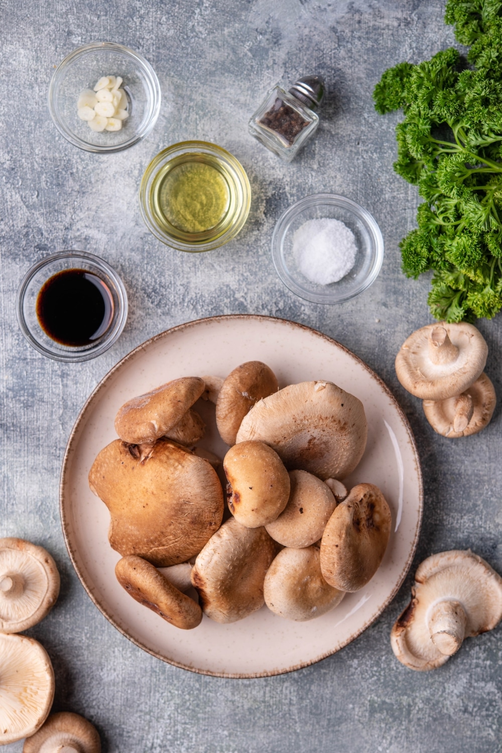 A large bowl of raw shiitake mushrooms and small glass bowls of salt, olive oil, soy sauce, and sliced garlic, plus a glass pepper shaker.