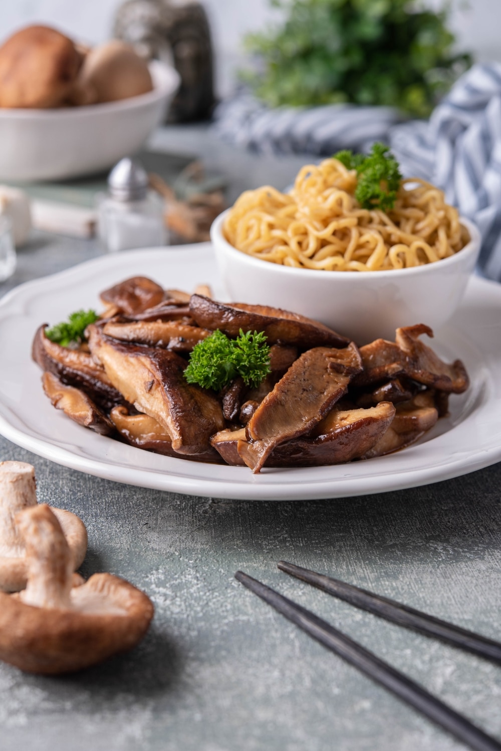 Sauteed shiitake mushrooms garnished with parsley on a white plate with a small bowl of noodles. Next to the plate is a pair of black chopsticks and behind it is a bowl of raw mushrooms.