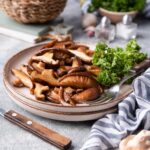 A plate of sauteed shiitake mushrooms with curly parsley on the side. A fork is on the plate and behind it is a basket of fresh mushrooms and a small bowl of parsley.