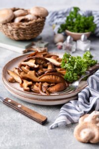 A plate of sauteed shiitake mushrooms with curly parsley on the side. A fork is on the plate and behind it is a basket of fresh mushrooms and a small bowl of parsley.