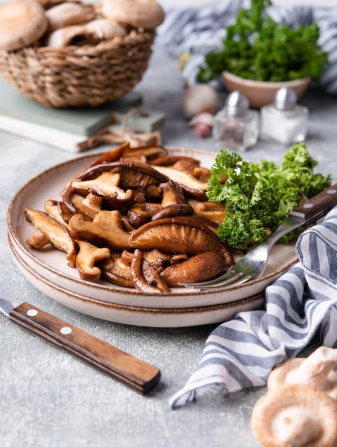 A plate of sauteed shiitake mushrooms with curly parsley on the side. A fork is on the plate and behind it is a basket of fresh mushrooms and a small bowl of parsley.