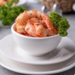 A small white bowl filled with sauteed shrimp with a piece of curly parsley on the side for garnish. In the back is a basket of lemons, a bunch of fresh curly parsley, and a plate of sauteed shrimp on pasta.