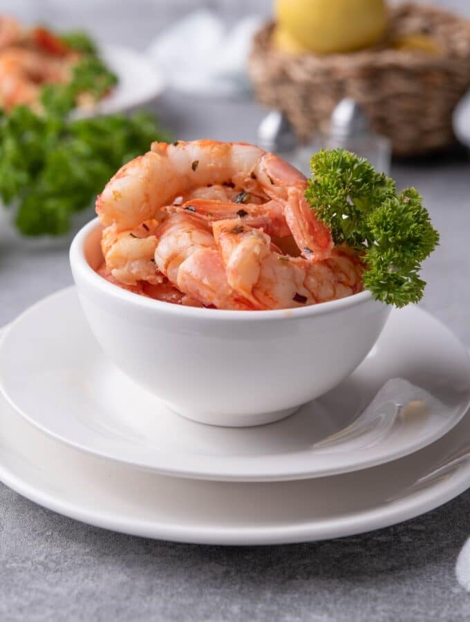 A small white bowl filled with sauteed shrimp with a piece of curly parsley on the side for garnish. In the back is a basket of lemons, a bunch of fresh curly parsley, and a plate of sauteed shrimp on pasta.