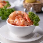 A small white bowl filled with sauteed shrimp with a piece of curly parsley on the side for garnish. In the back is a basket of lemons, a bunch of fresh curly parsley, and a plate of sauteed shrimp on pasta.