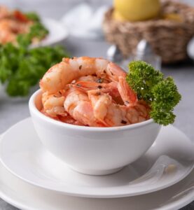 A small white bowl filled with sauteed shrimp with a piece of curly parsley on the side for garnish. In the back is a basket of lemons, a bunch of fresh curly parsley, and a plate of sauteed shrimp on pasta.