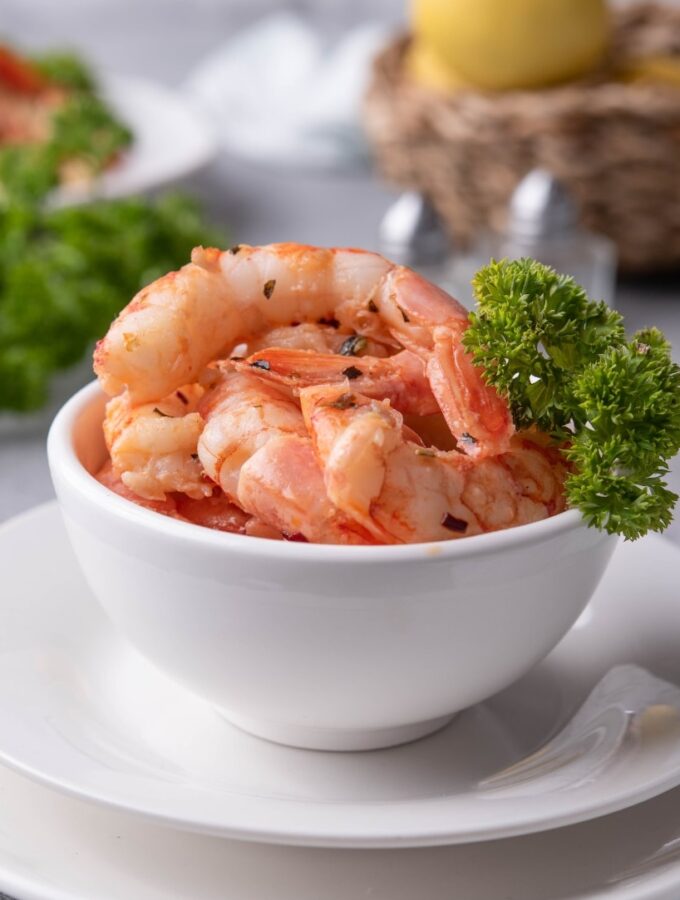 A small white bowl filled with sauteed shrimp with a piece of curly parsley on the side for garnish. In the back is a basket of lemons, a bunch of fresh curly parsley, and a plate of sauteed shrimp on pasta.