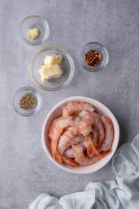 A white bowl with tail-on shrimp that's been deshelled and deveined, a small glass bowl of softened butter, and smaller glass bowls of minced garlic, chili flakes, and dried Italian herbs.