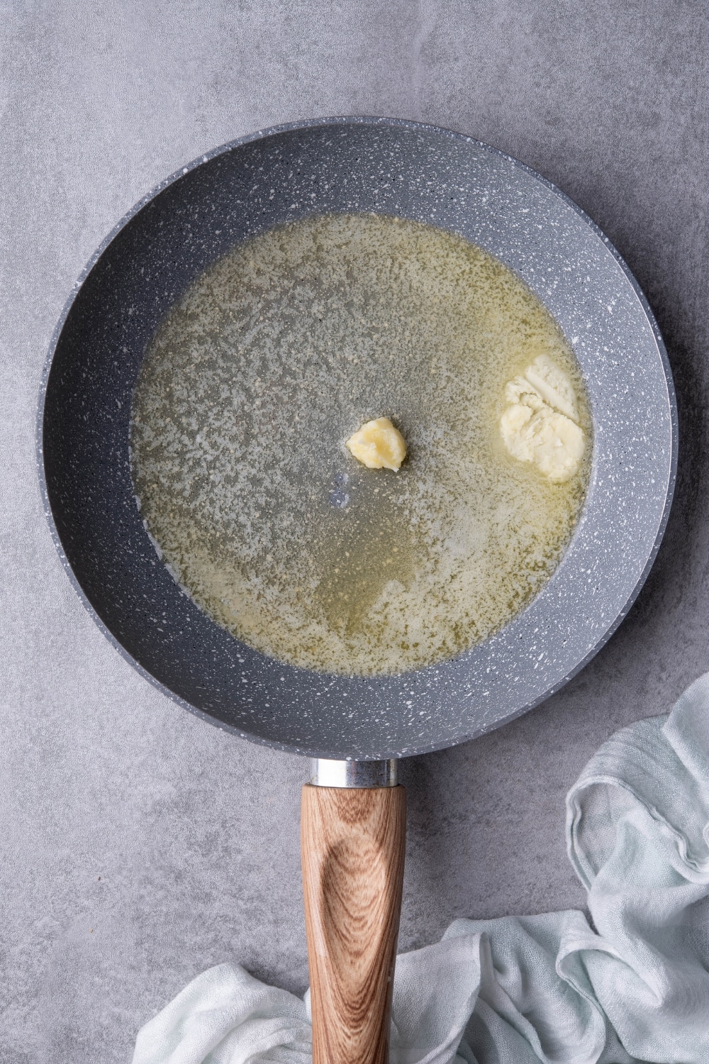 Butter melting in a speckled grey pan with a wooden handle. The pan is on a grey countertop.