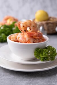 A small white bowl filled with sauteed shrimp. The bowl is resting on a white plate with a piece of curly parsley on the side for garnish. In the back is a basket of lemons, a bunch of fresh curly parsley, and a plate of sauteed shrimp over pasta.
