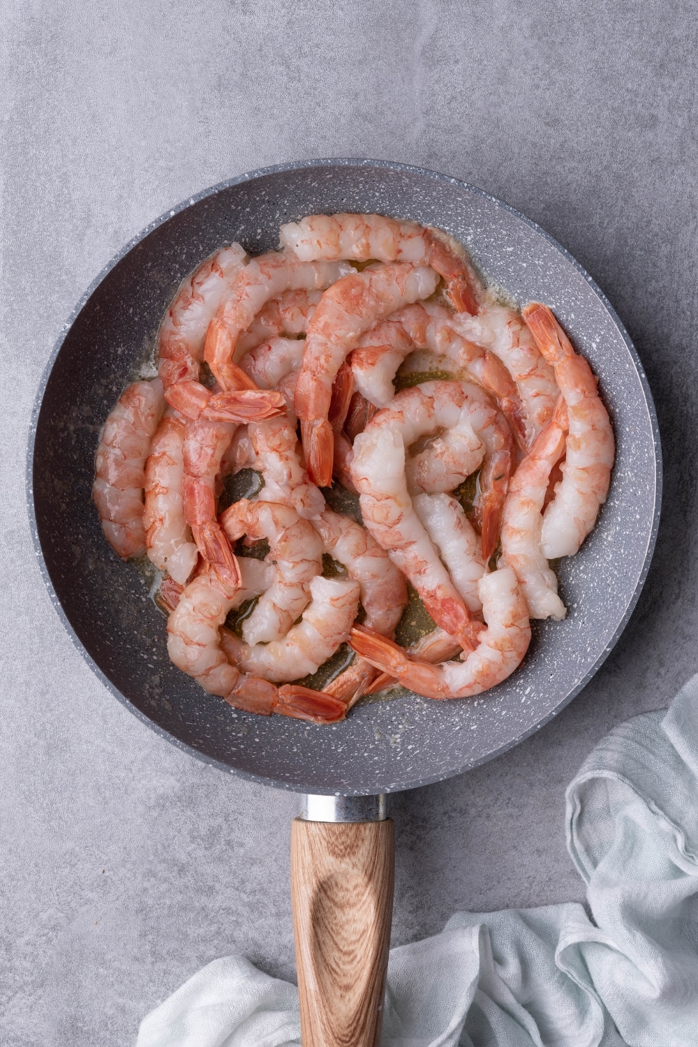 Partially cooked tail-on shrimp sauteing in melted butter in a speckled grey pan. The pan has a wooden handle and is resting on a grey countertop.