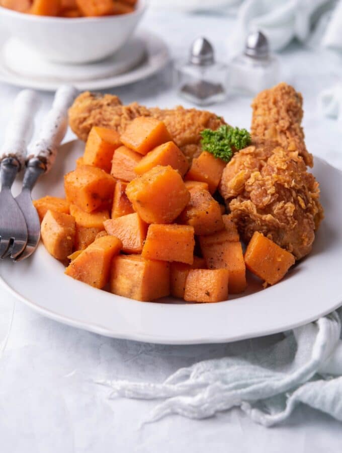 Sauteed sweet potatoes on a plate served with two breaded chicken drumsticks and a pair of forks with white handles. Behind the plate is a pair of salt and pepper shakers and a small bowl with more sauteed sweet potatoes.