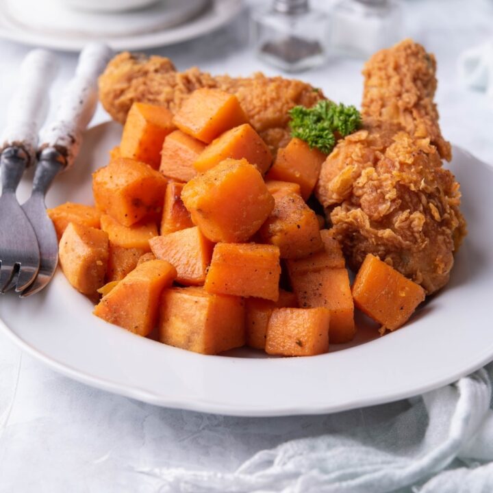 Sauteed sweet potatoes on a plate served with two breaded chicken drumsticks and a pair of forks with white handles. Behind the plate is a pair of salt and pepper shakers and a small bowl with more sauteed sweet potatoes.
