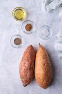 Two sweet potatoes and small glass bowls of olive oil, cumin, salt, and pepper.