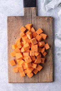 Cubed sweet potatoes on a wooden cutting board with a black handle. The cutting board is on a grey countertop next to a tea towel.