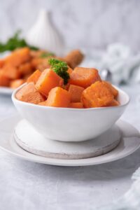 Sauteed sweet potato cubes garnished with parsley in a small white bowl over a white plate. Behind the bowl is a plate of more sauteed sweet potatoes served with fried chicken.