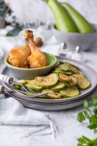 Sauteed zucchini on stacked pair of blue grey plates with a small bowl of fried chicken drumsticks on the side. Two forks are resting on the plate and in the back are salt and peppers shakers and a bowl of whole zucchinis