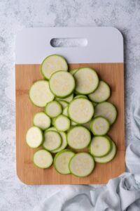 Raw zucchini slices on a wooden cutting board with a white handle. The cutting board is on a grey countertop and a tea towel is on the side.