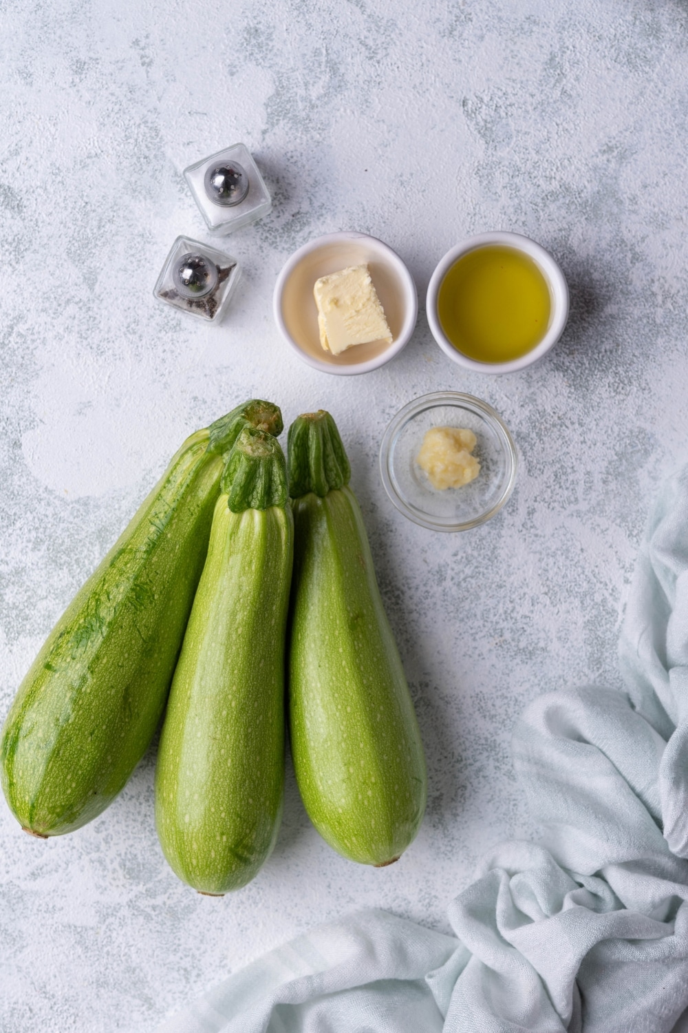 Three medium sized zucchini, salt and pepper shakers, two small ceramic ramekins of olive oil and butter, and a small glass bowl of garlic paste.