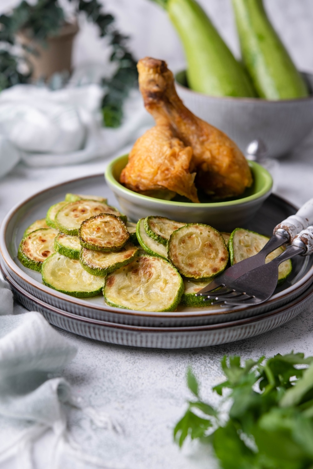 Sauteed zucchini slices on stacked blue grey plates with a small bowl of two fried chicken drumsticks on the side. Two forks are also on the plate and in the back is a bowl of whole zucchinis.