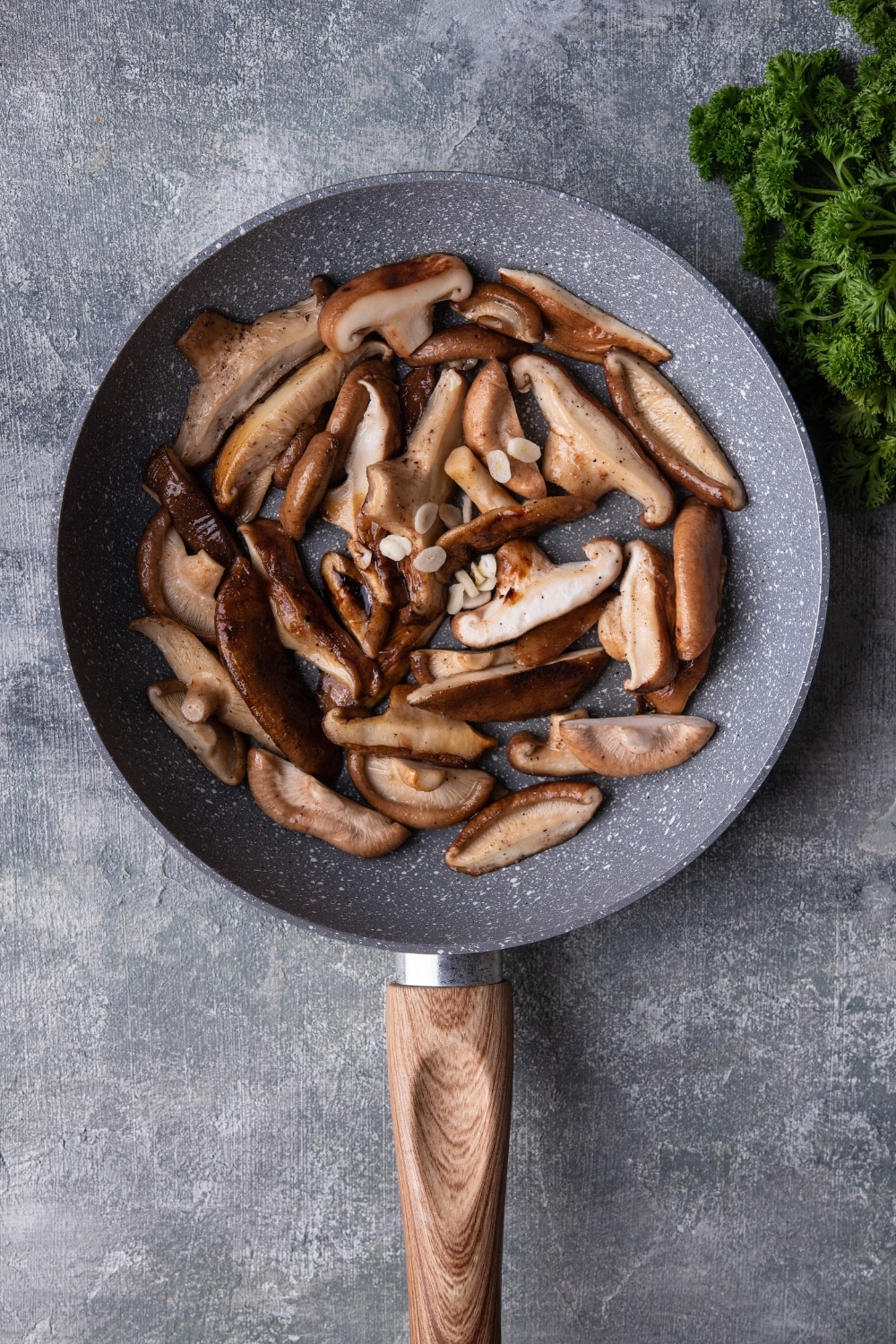 Sauteed shiitake mushrooms in a grey speckled pan with a wooden handle. The pan is on a grey countertop next to a bunch of fresh parsley.