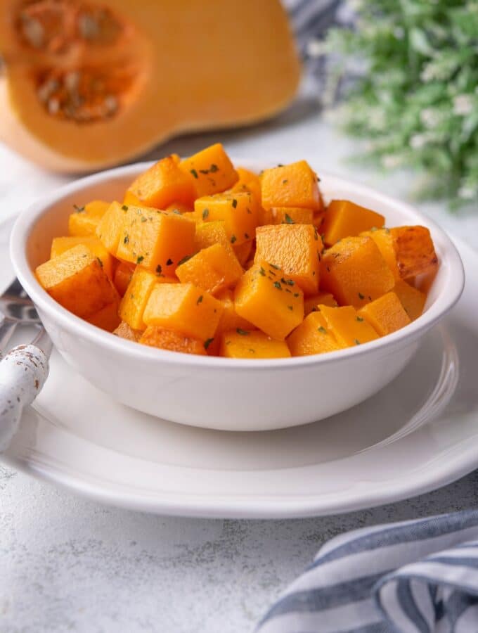 Sauteed butternut squash cubes garnished with herbs in a white bowl. The bowl is set atop a white plate next to a metal fork and knife with white handles. In the back is a halved butternut squash and a striped tea towel.