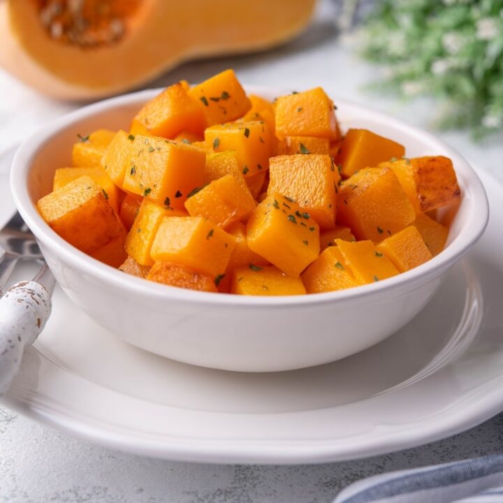 Sauteed butternut squash cubes garnished with herbs in a white bowl. The bowl is set atop a white plate next to a metal fork and knife with white handles. In the back is a halved butternut squash and a striped tea towel.