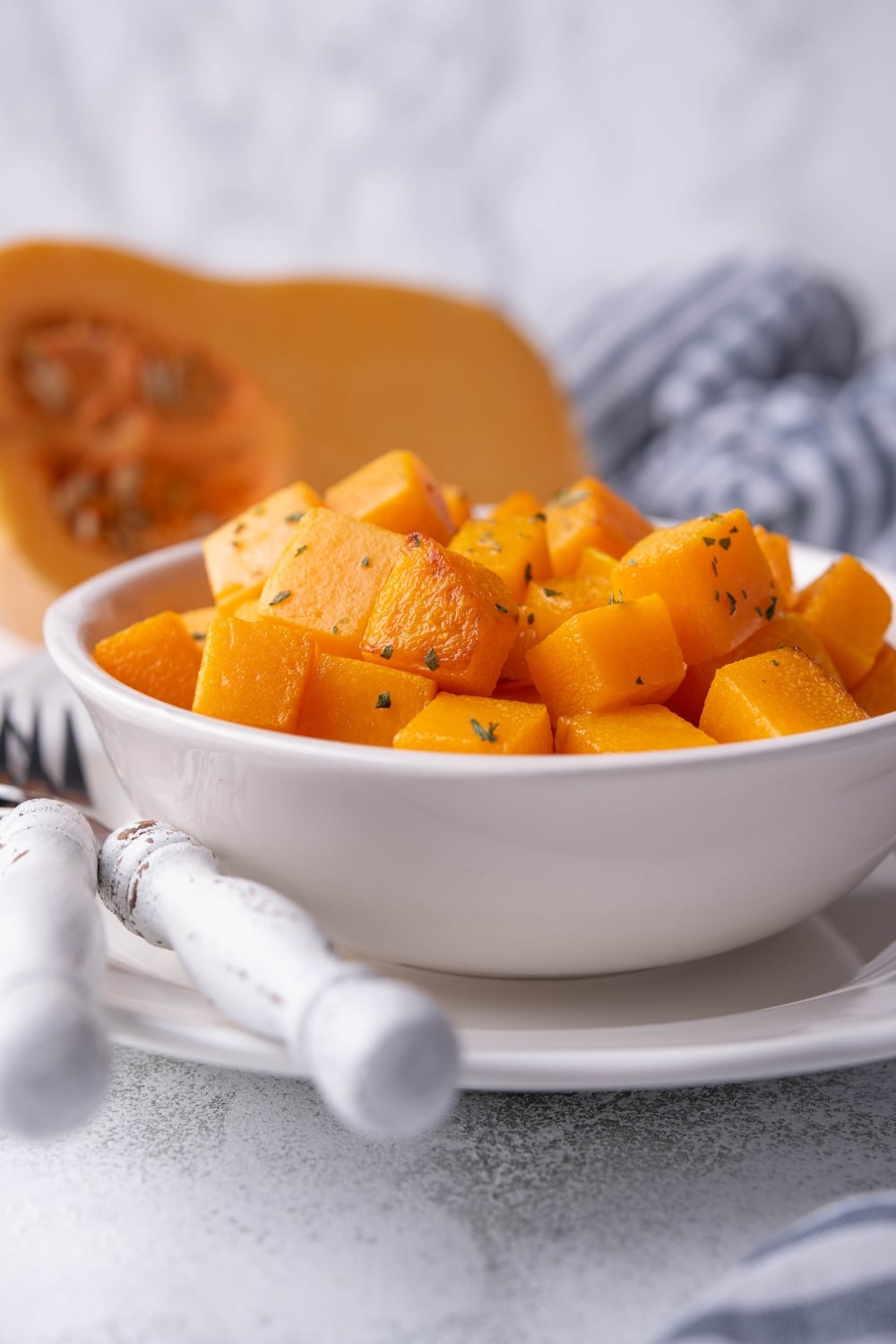 A closer look at the sauteed butternut squash cubes garnished with herbs in a white bowl. The bowl is on top of a white plate next to a fork and knife set with white handles.A halved butternut squash and a striped tea towel are in the back.