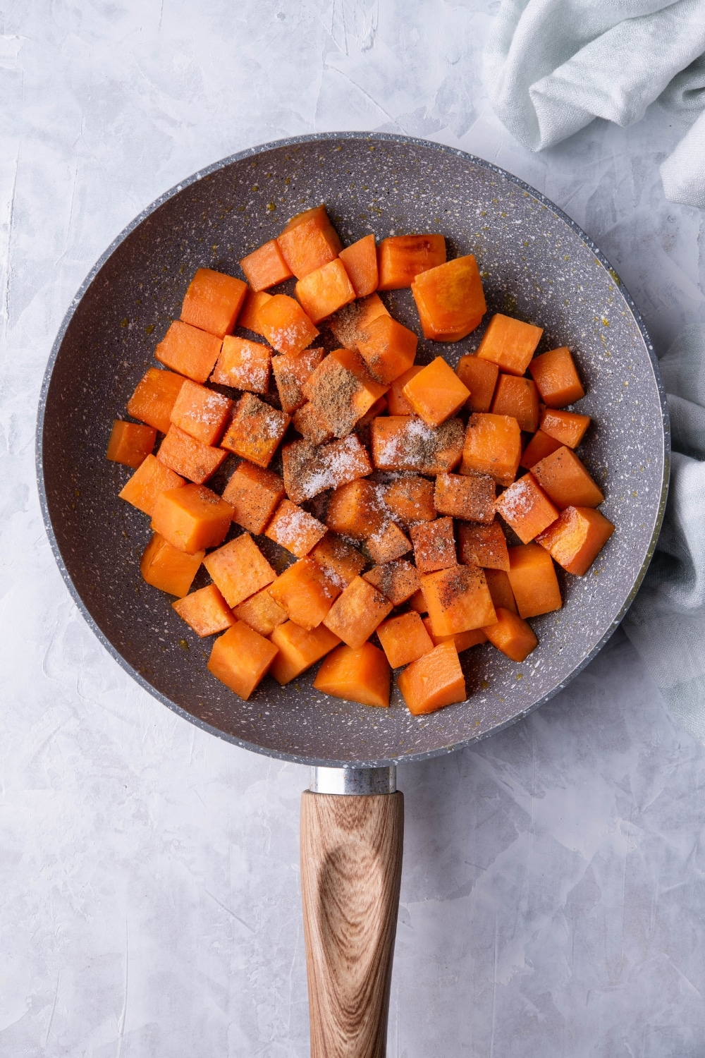 Diced sweet potatoes covered with spices in a grey speckled skillet with a wooden handle. The skillet is on a grey countertop next to a tea towel.