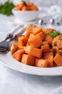 Close up of sauteed sweet potatoes on a plate with a pair of forks. Part of a pair of breaded chicken drumsticks can be seen on the same plate. Behind is a pair of salt and pepper shakers and a small bowl with more sauteed sweet potatoes.