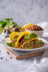 Ground turkey tacos with avocados, mixed greens, and cilantro on a plate with a lime wedge. Behind is a bowl of seasoned ground turkey meat and a sliced avocado.