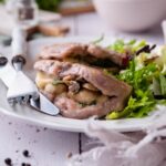 Seared pork chops stuffed with swiss cheese and mushrooms on a white plate with garden salad. A fork and knife are on the plate and a bowl of salad can be seen in the back.